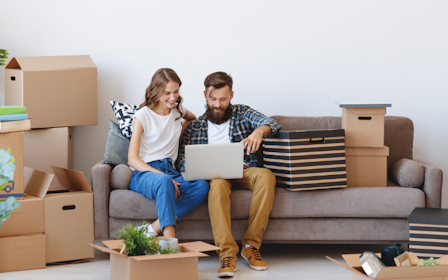 A man and a woman sitting on a couch with boxes and a laptop.